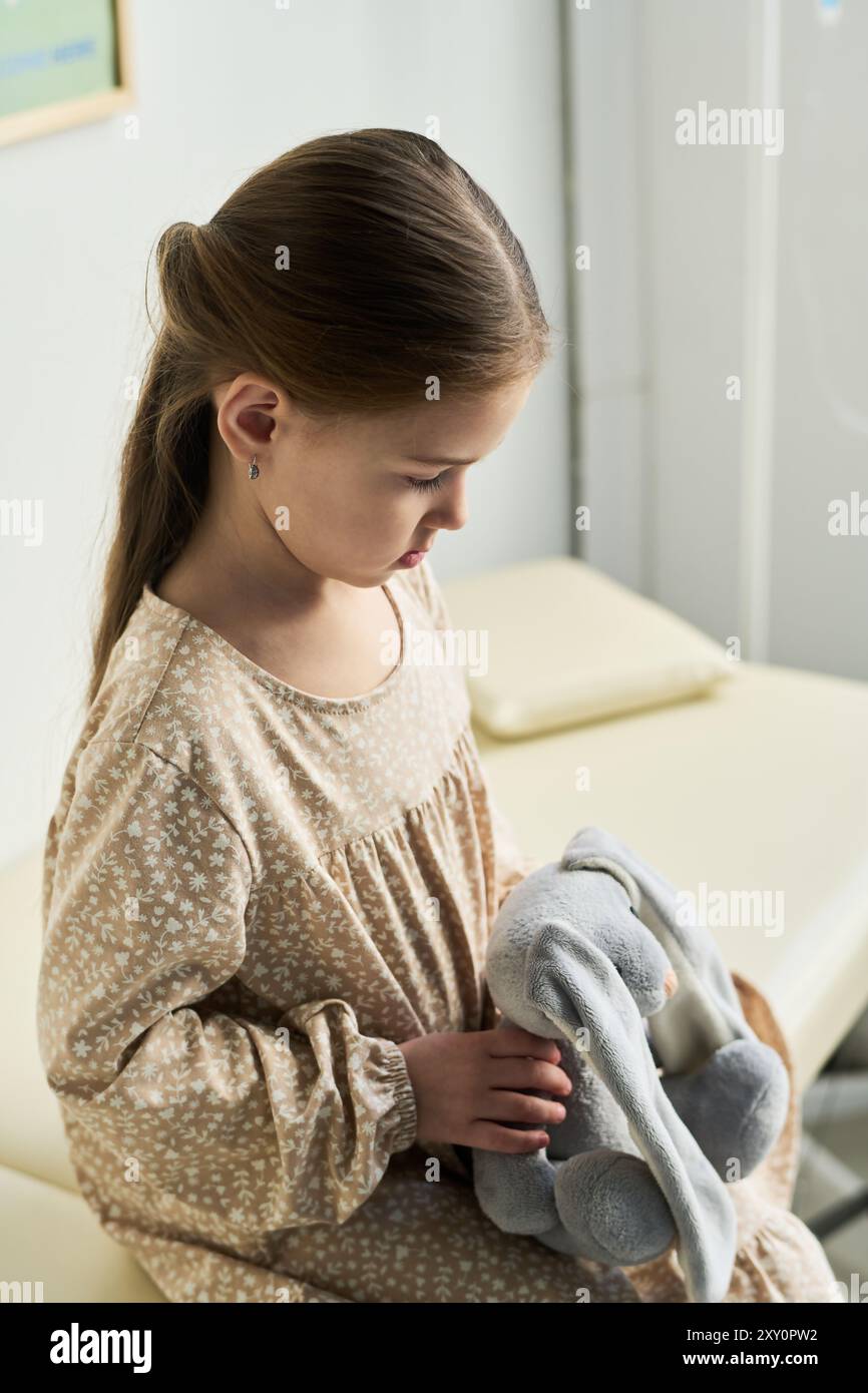 Brown-haired girl sitting indoors holding stuffed elephant toy, wearing beige long-sleeved dress, looking down with a reflective expression Stock Photo