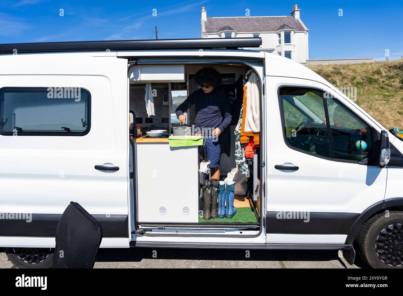 A young man enjoys the convenience of his camper van by preparing lunch inside the compact, well-equipped kitchen on a sunny summer day, surrounded by Stock Photo