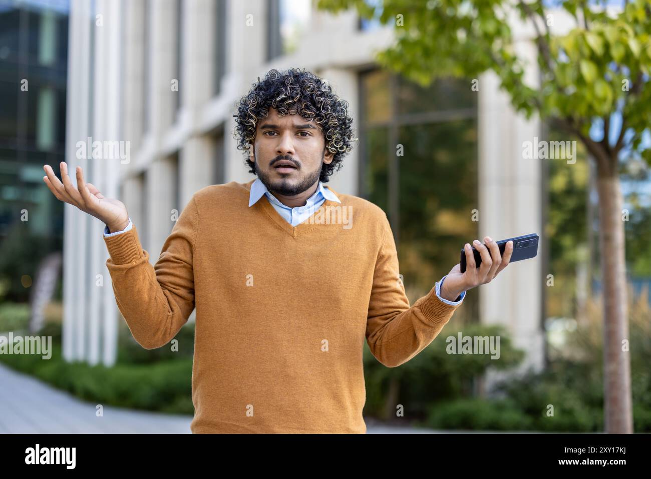 Young man with curly hair shrugs while holding smartphone, expressing confusion or uncertainty. Stock Photo