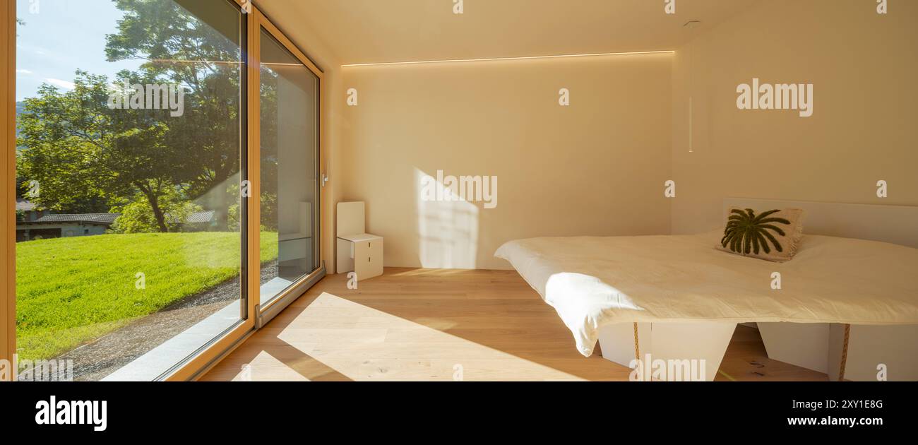 Front view of a bedroom with cardboard home staging furniture, a very bright window overlooking nature, green lawn and trees. No one inside. Stock Photo