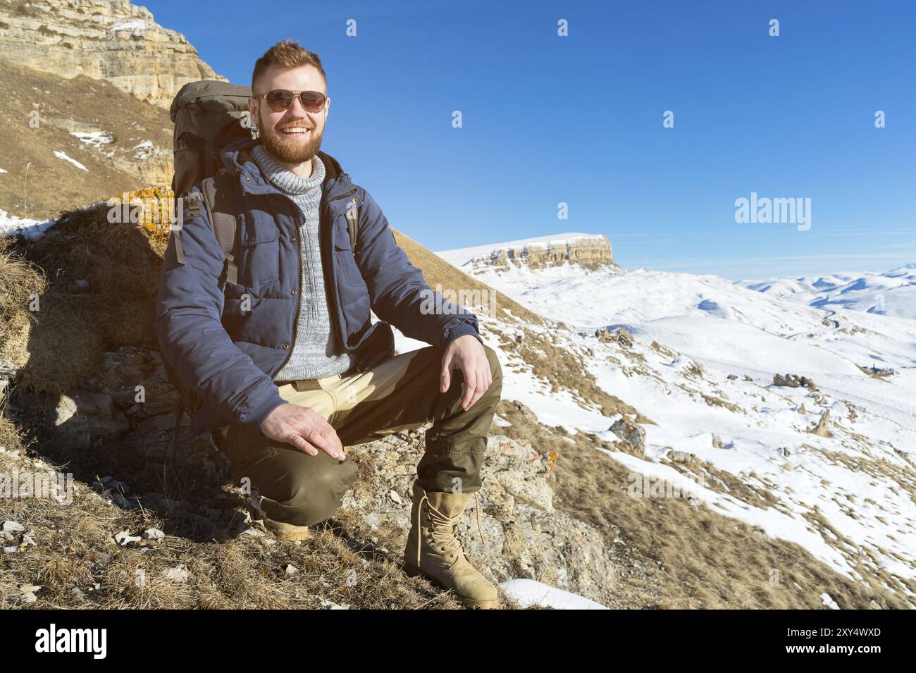 Portrait of happy laughing Hipster traveler with beard in sunglasses is sitting on nature. A man hiking in the mountains with a backpack and Scandinav Stock Photo