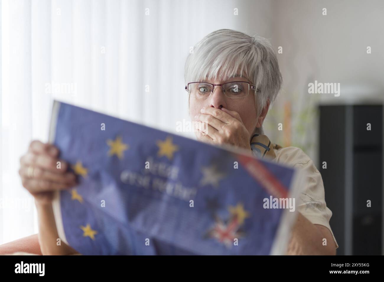 An elderly lady reads the news about Brexit and puts her hand over her mouth in horror Stock Photo