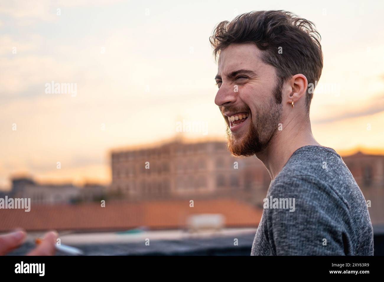 Candid portrait of a cheerful young bearded man laughing heartily on a city rooftop at sunset. Natural, genuine emotion captured in warm evening light Stock Photo