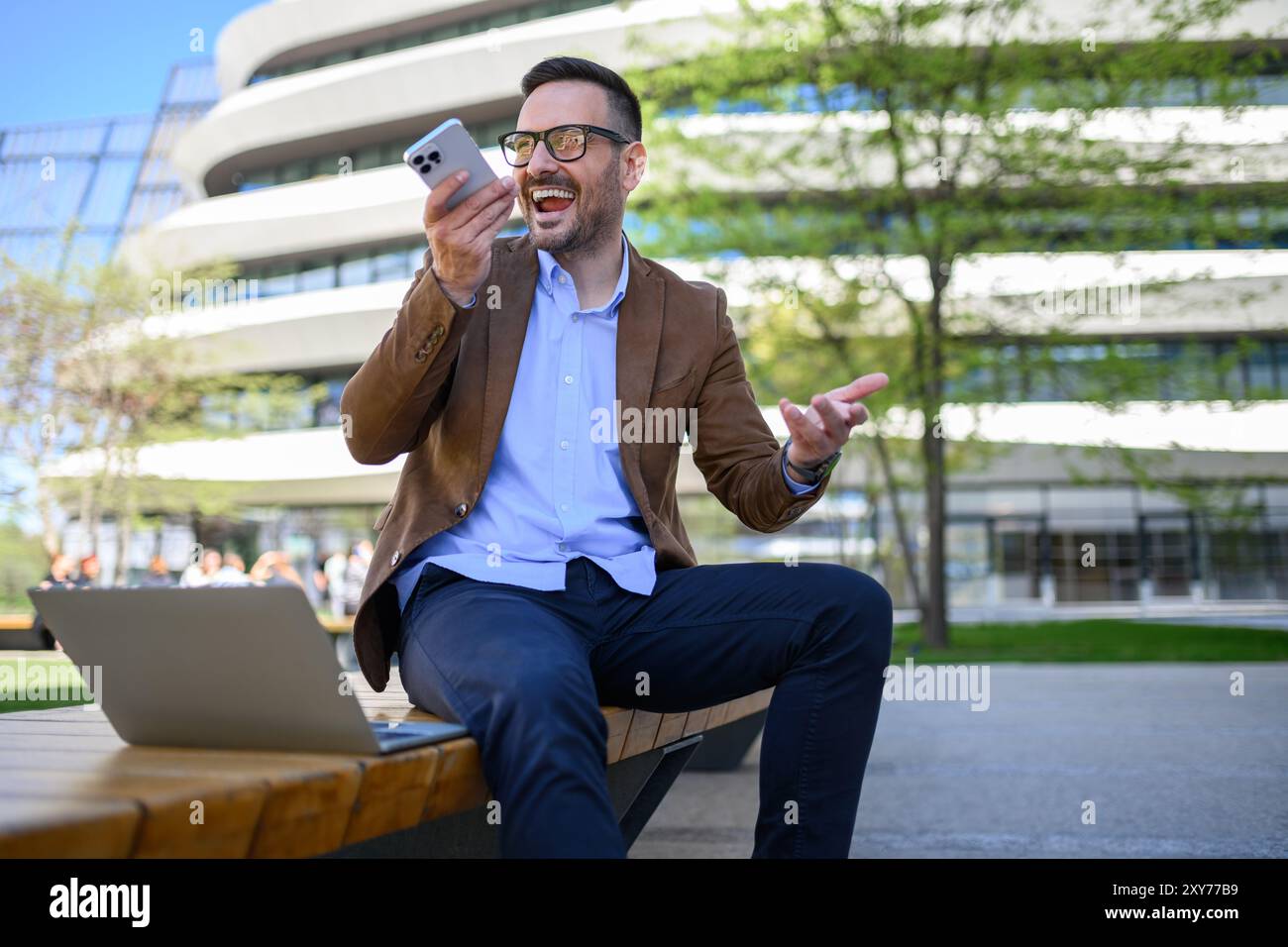 Cheerful male professional talking over phone speaker and working over laptop on bench in city Stock Photo