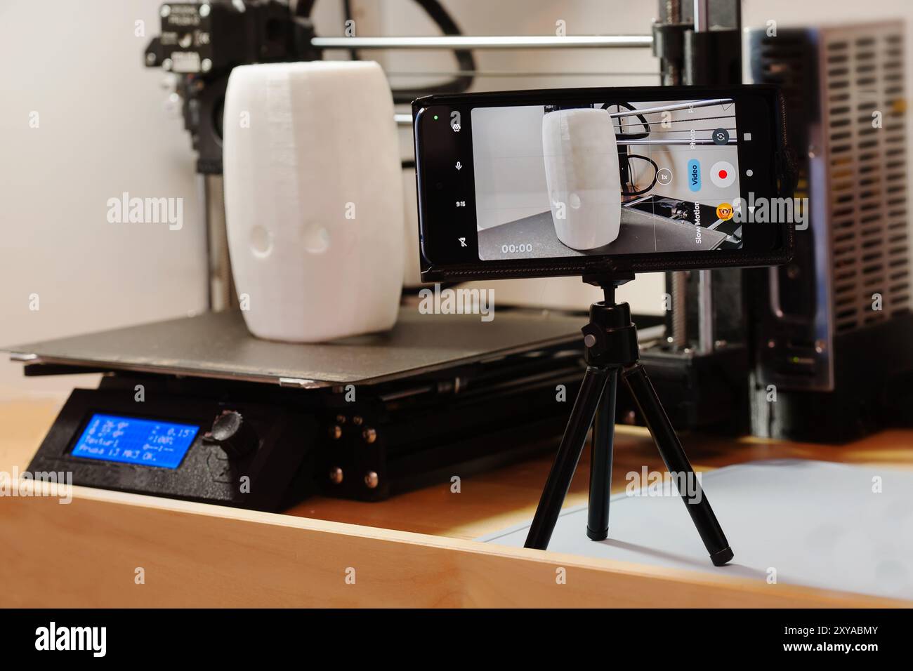 Phone mounted on a small tripod being used to record a successful 3d print of a tall white object sitting on a 3d printer's printbed. Stock Photo
