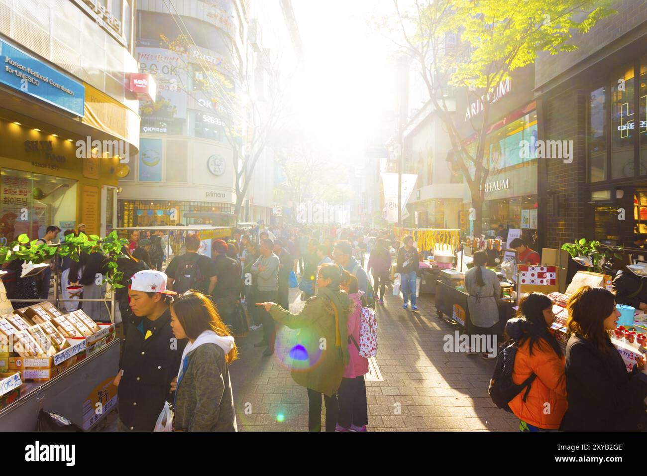 Seoul, South Korea, April 17, 2015: Sun backlights tourists walking down busy Myeongdong pedestrian shopping street with commercialism of stores, sign Stock Photo