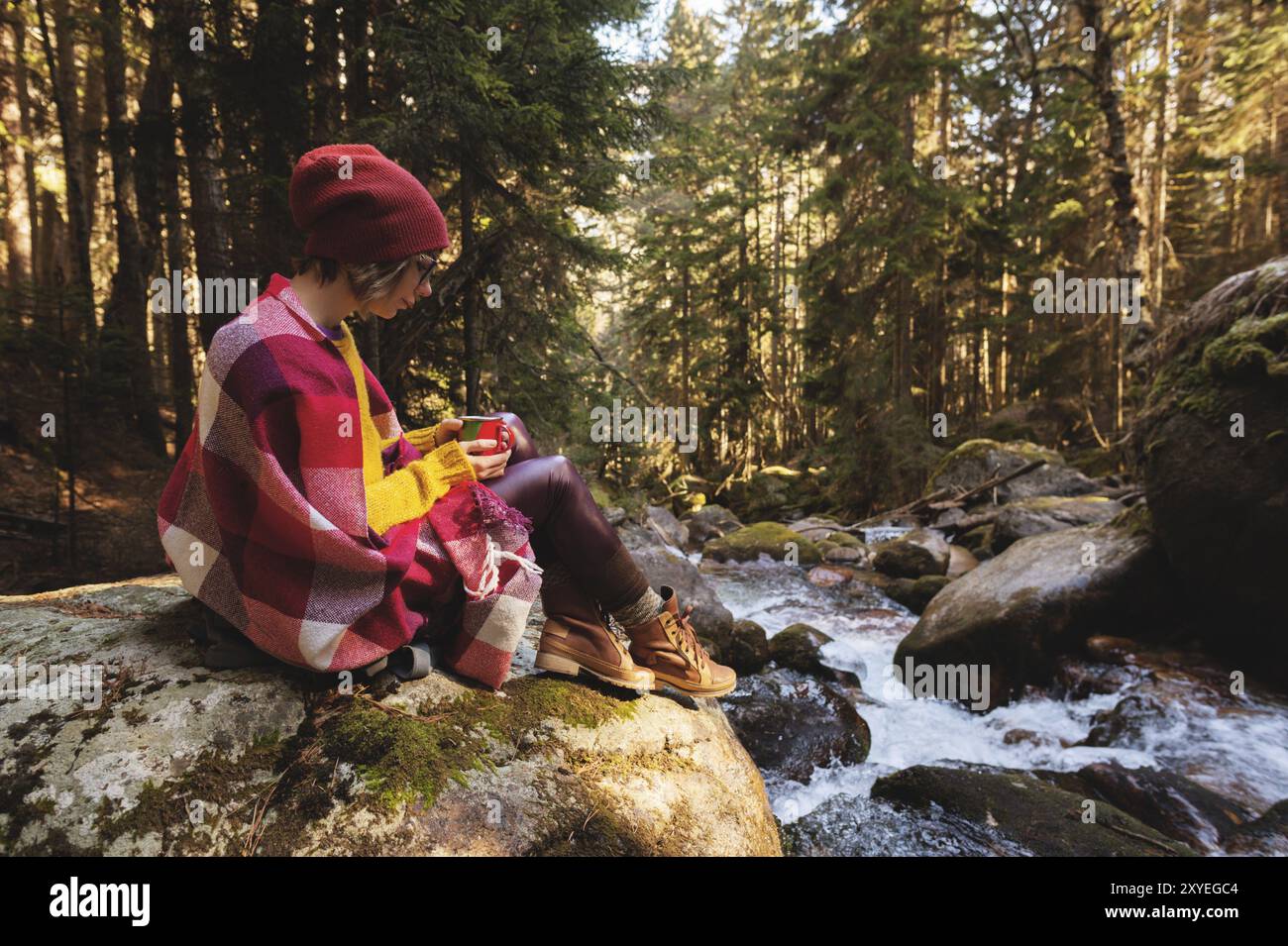 A pretty hipster girl with a plaid plaid on her shoulders in a hat and a yellow sweater and glasses with a mug of coffee in her hands looks at a mount Stock Photo