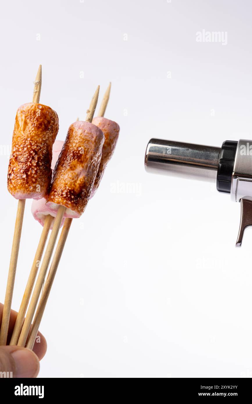 Horizontal image showing toasted marshmallows on skewers being roasted with a torch. The hand holding the torch is on the left, set against a clean wh Stock Photo