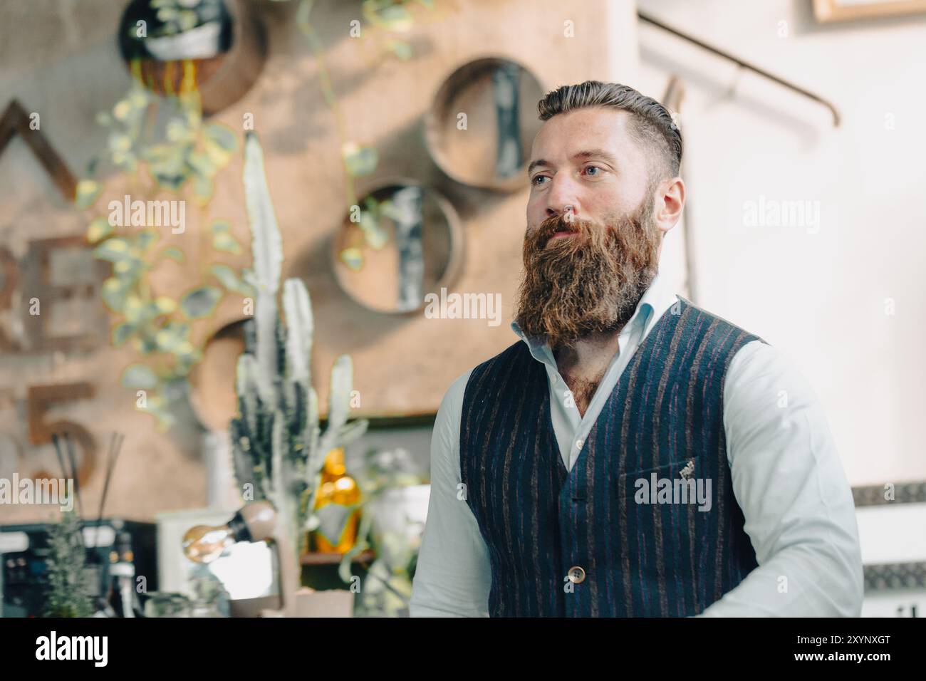 Bearded businessman wearing a vest and looking away while standing in a modern office with plants Stock Photo