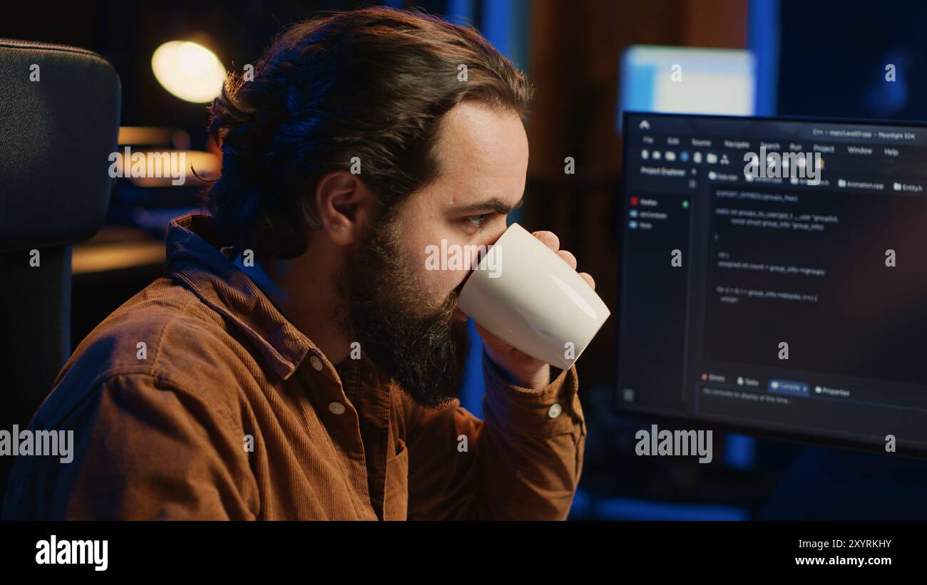 Focused programmer drinking coffee in apartment while solving database errors on computer. Remotely working IT admin concentrated on finalizing coding project in home office, camera A Stock Photo