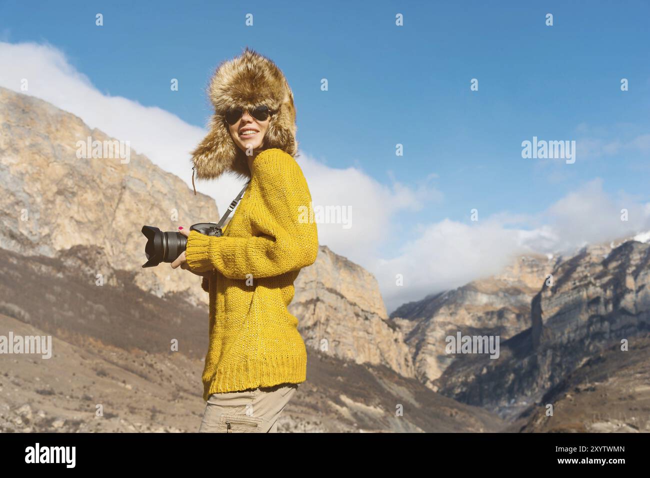 A girl photographer in sunglasses and a big fur hat and a yellow knitted sweater stands against the background of high rocks in the gorge with a camer Stock Photo