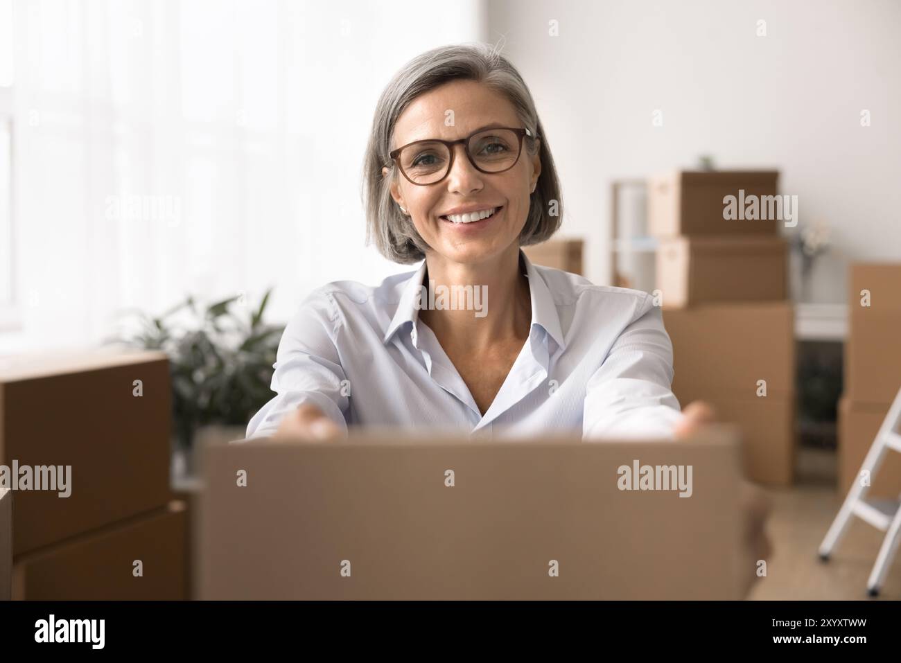 Smiling, professional middle-aged woman extending cardboard box towards viewer Stock Photo