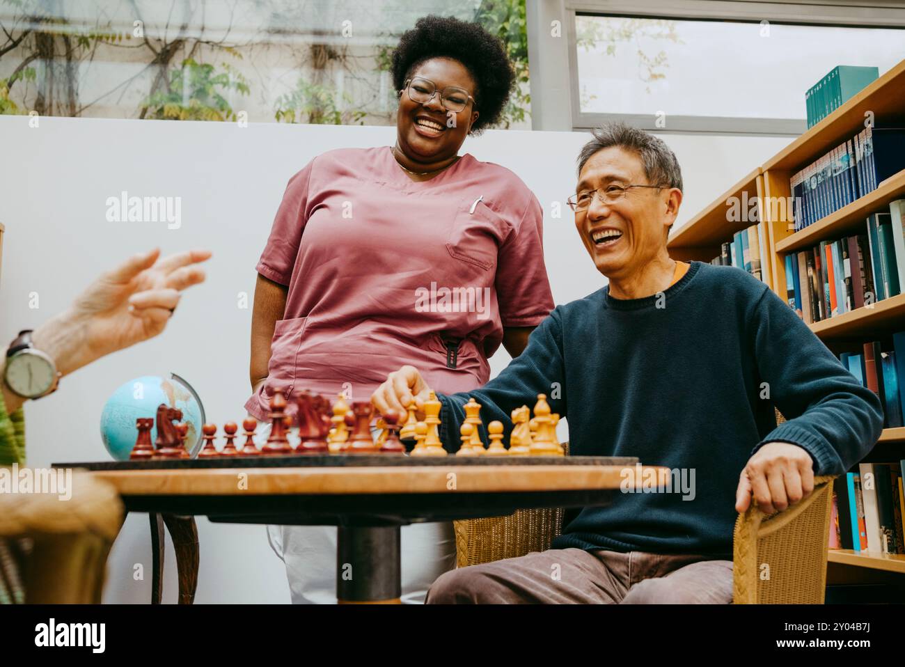 Happy female caregiver standing near senior man playing chess while sitting in nursing home Stock Photo