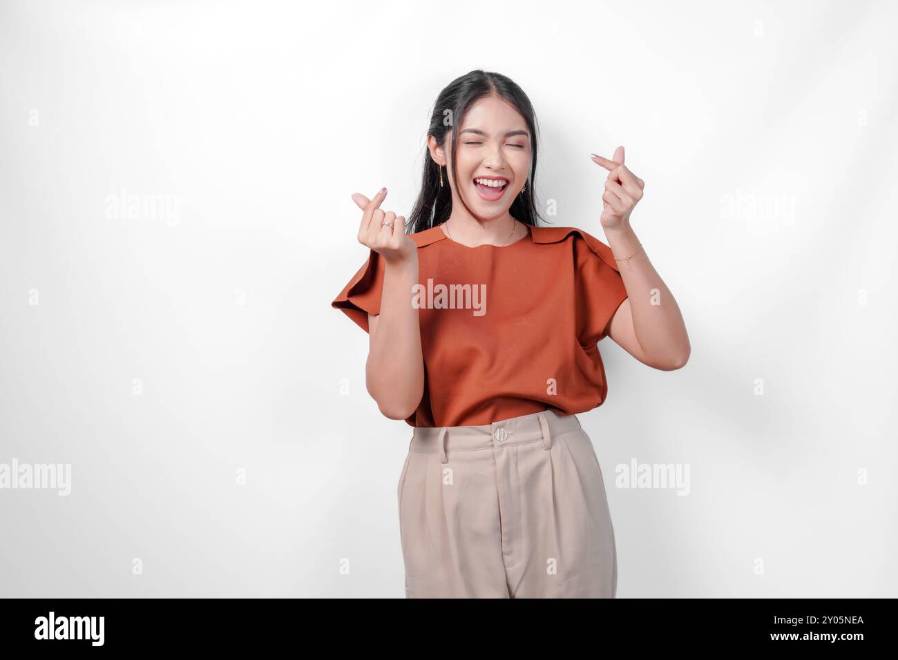 Lovely young Asian woman in a brown shirt is smiling and showing finger heart gesture to the camera, isolated by white background. Stock Photo