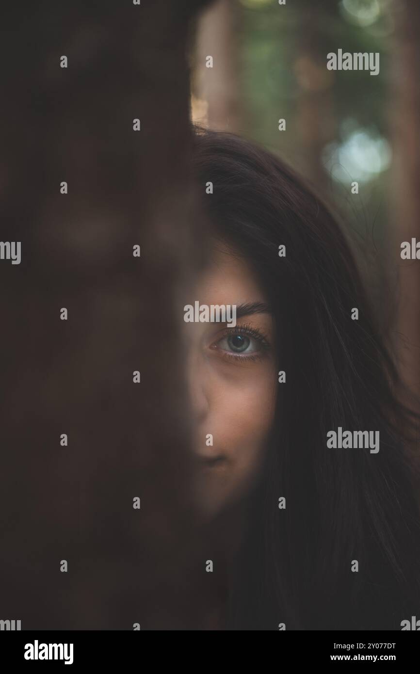 Enigmatic woman with captivating blue eyes peeks from behind a tree in a mystical forest. The image captures the essence of mystery, nature, and seren Stock Photo