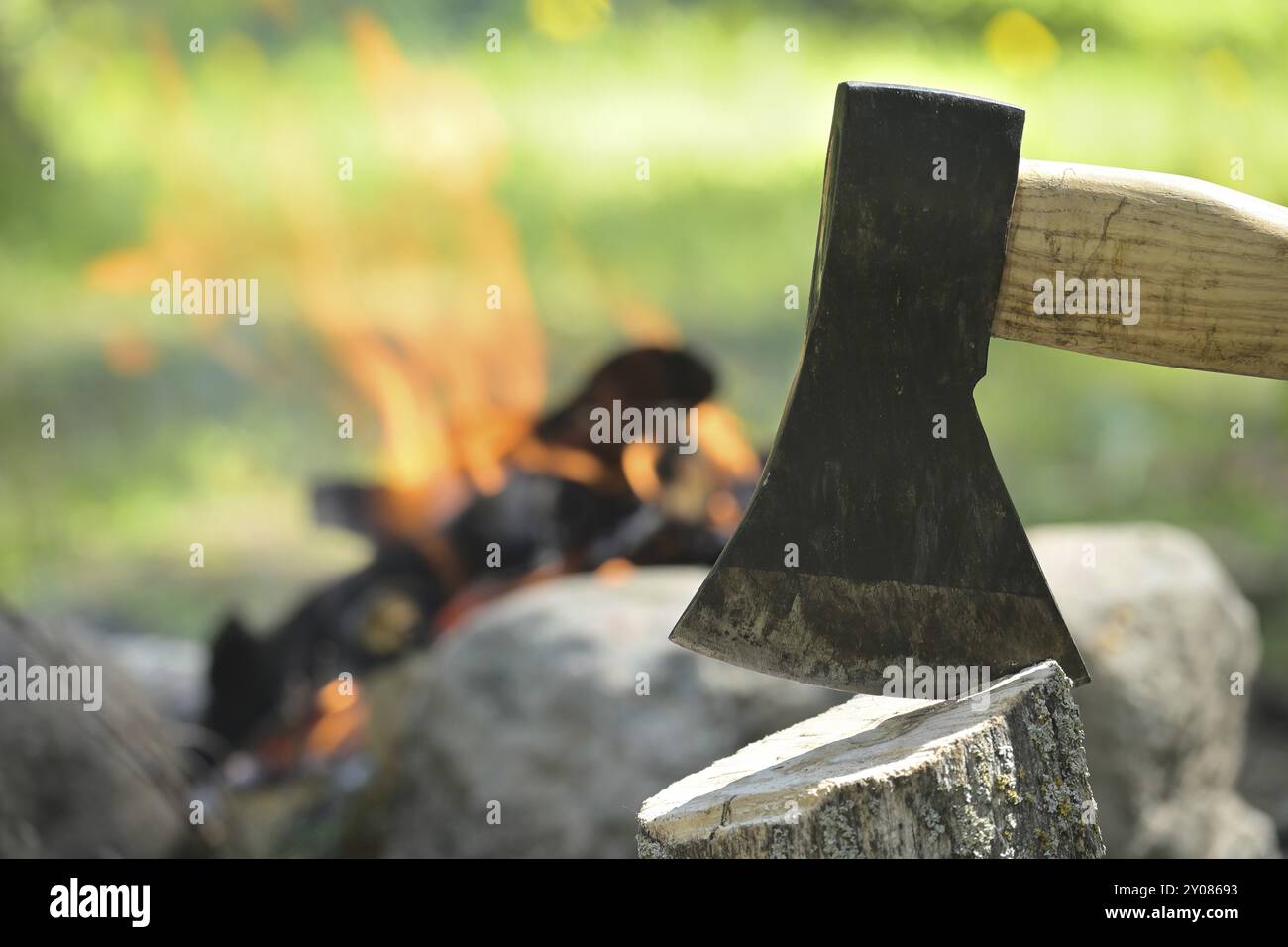Closeup view of axe with a wooden handle embedded in a log of wood, with a campfire in the background Stock Photo