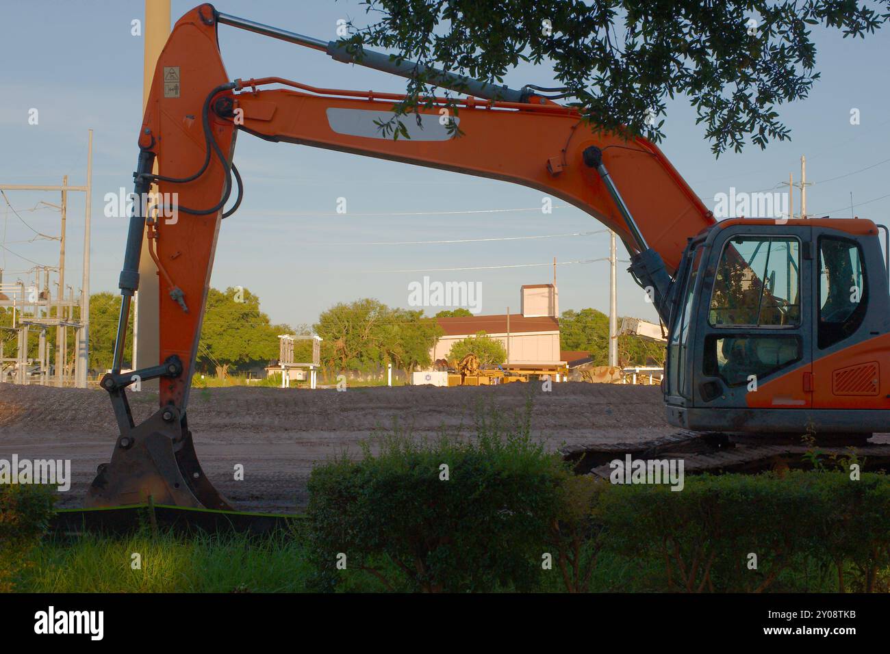 Wide framed view through orange bucket excavator machine at construction site of Electric Substation View over dirt pile with green grass in front Stock Photo