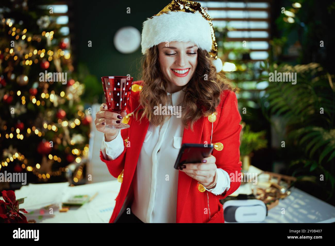 Christmas time. smiling stylish middle aged business woman in santa hat and red jacket with wineglass sending text message using smartphone in modern Stock Photo