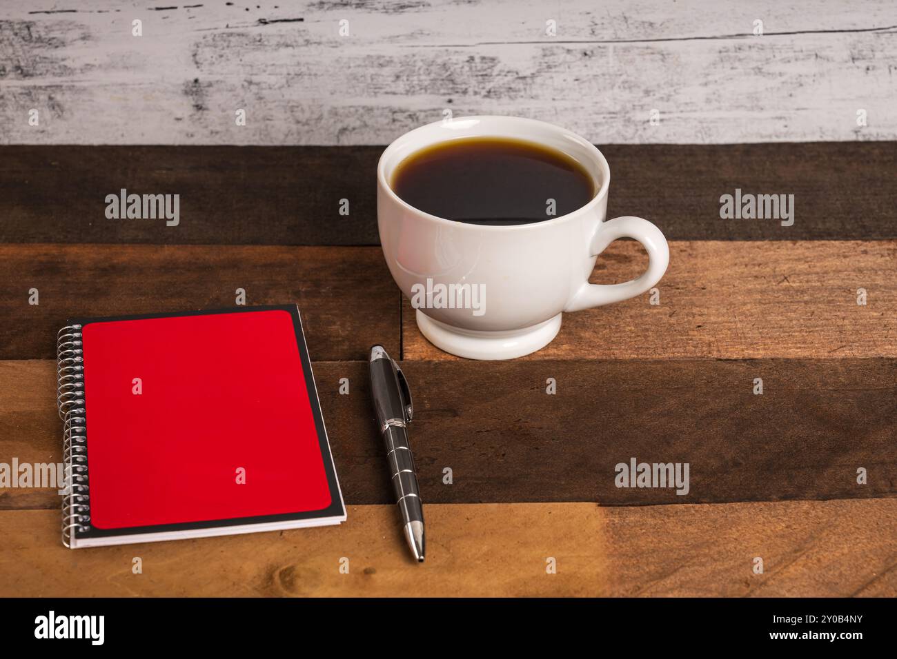 Coffee in white porcelain cup on rustic wood table with red and black covered spiral notebook and ball point pen Stock Photo