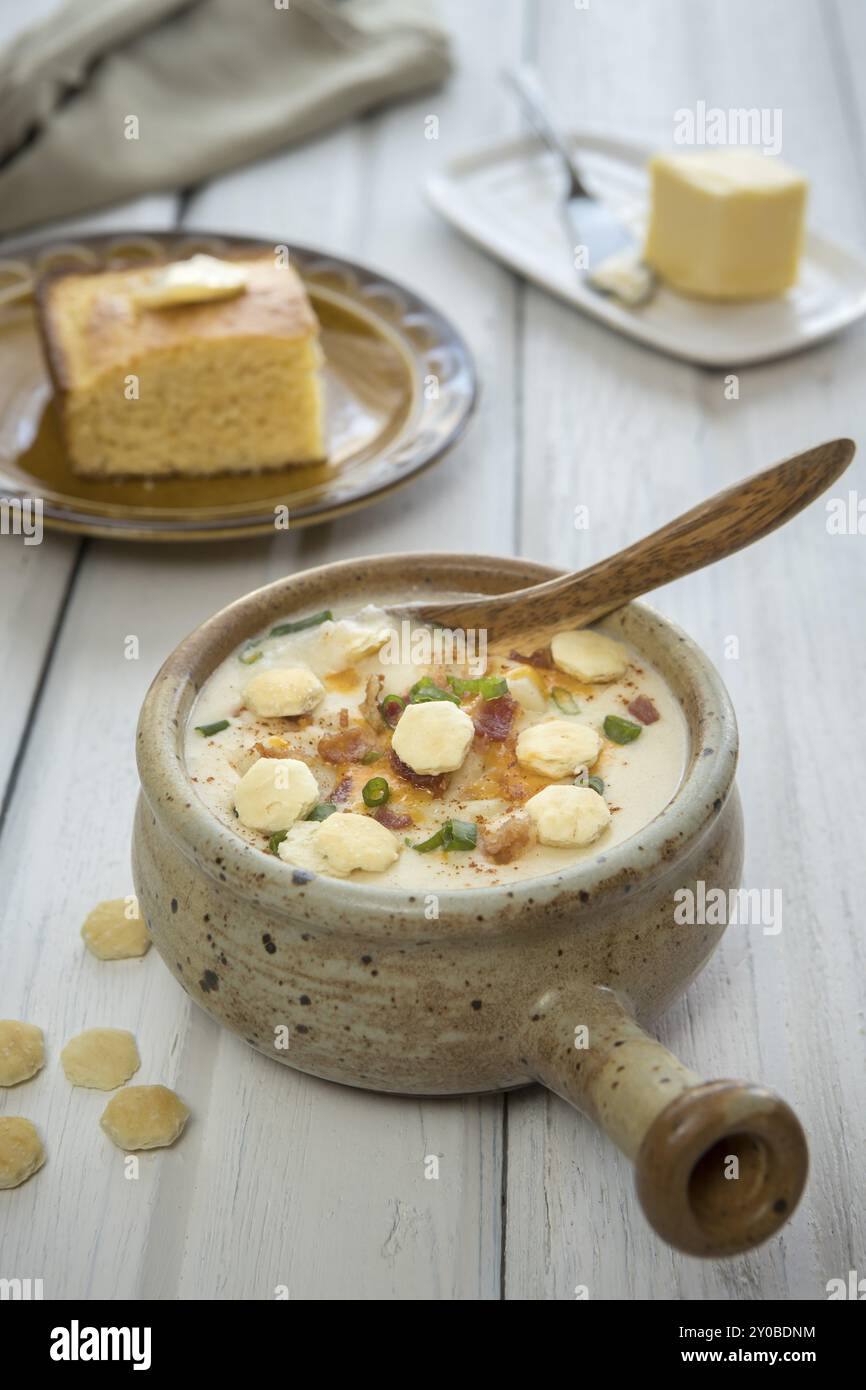 A bowl of homemade creamy potato and bacon soup topped with cheddar cheese, green onions, and oyster crackers with a side of cornbread Stock Photo