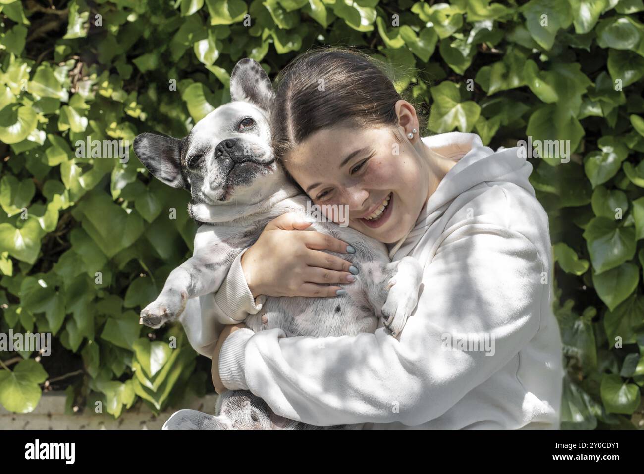 A girl in a white sweatshirt smiling and hugging a French Bulldog in a garden with lush green leaves in the background Stock Photo
