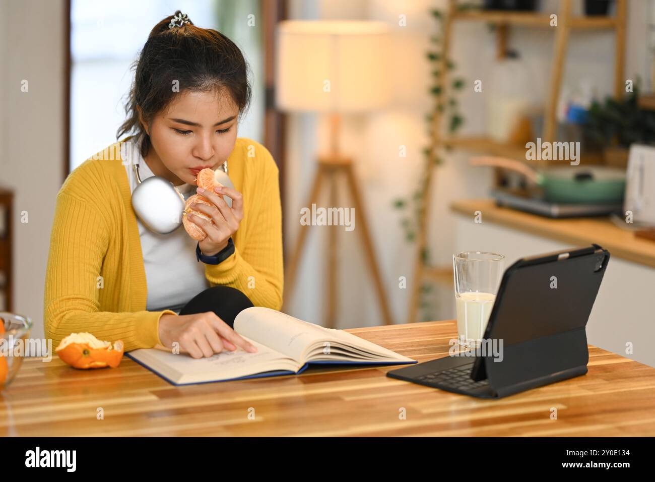 Beautiful young asian woman reading a book eating an orange at kitchen ...