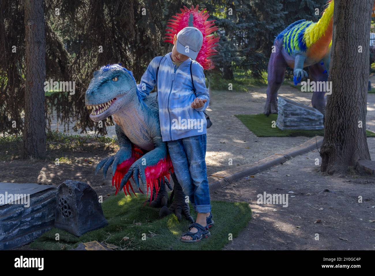 Boy hugs real-sized dinosaur with colored feathers at an amusement park. Stock Photo