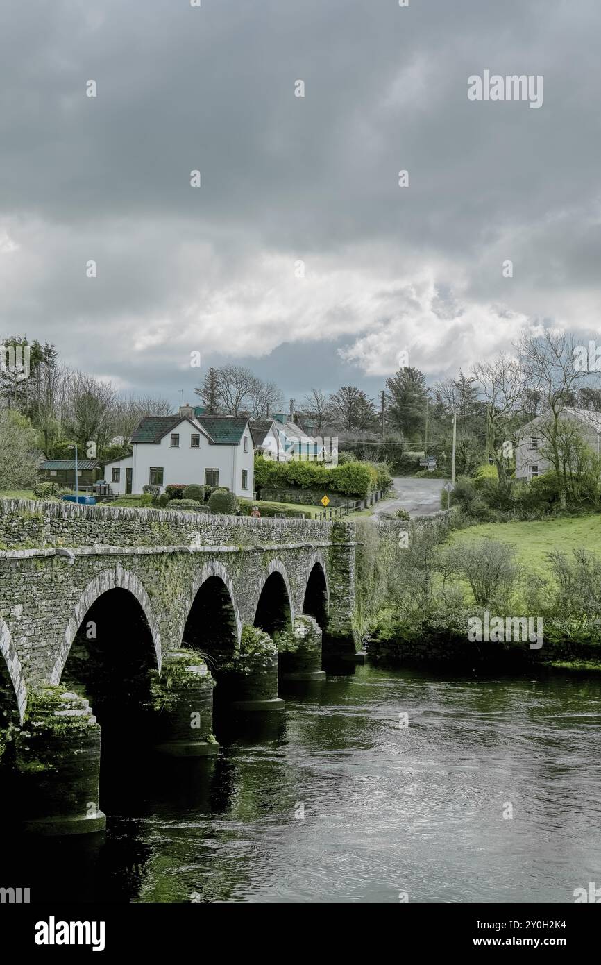 A stone bridge with multiple arches crosses a river, with a road leading to a small village on the other side. The sky is overcast. Bridge over the Ri Stock Photo