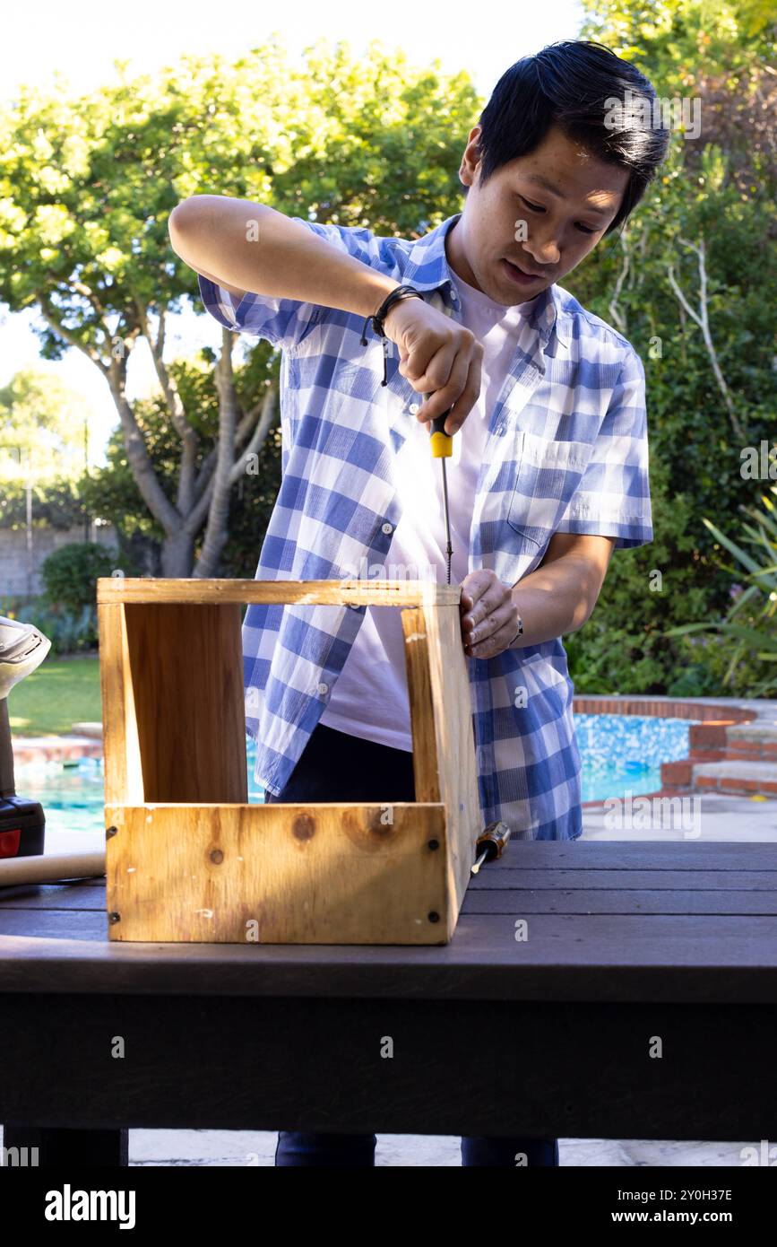 Using screwdriver, asian man assembling wooden box on outdoor table in backyard Stock Photo