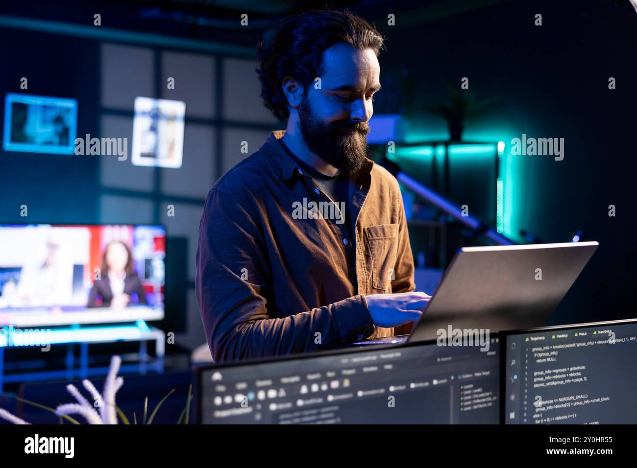 Smiling technician doing software debugging using laptop, checking glitches in lines of code displayed on PC monitors. Happy man walking in apartment fixing coding issues using notebook Stock Photo