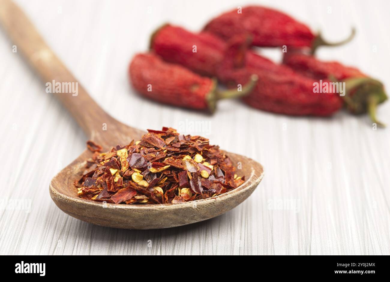 A close up photo of red pepper flakes on a wooden spoon and whole red peppers in the background Stock Photo