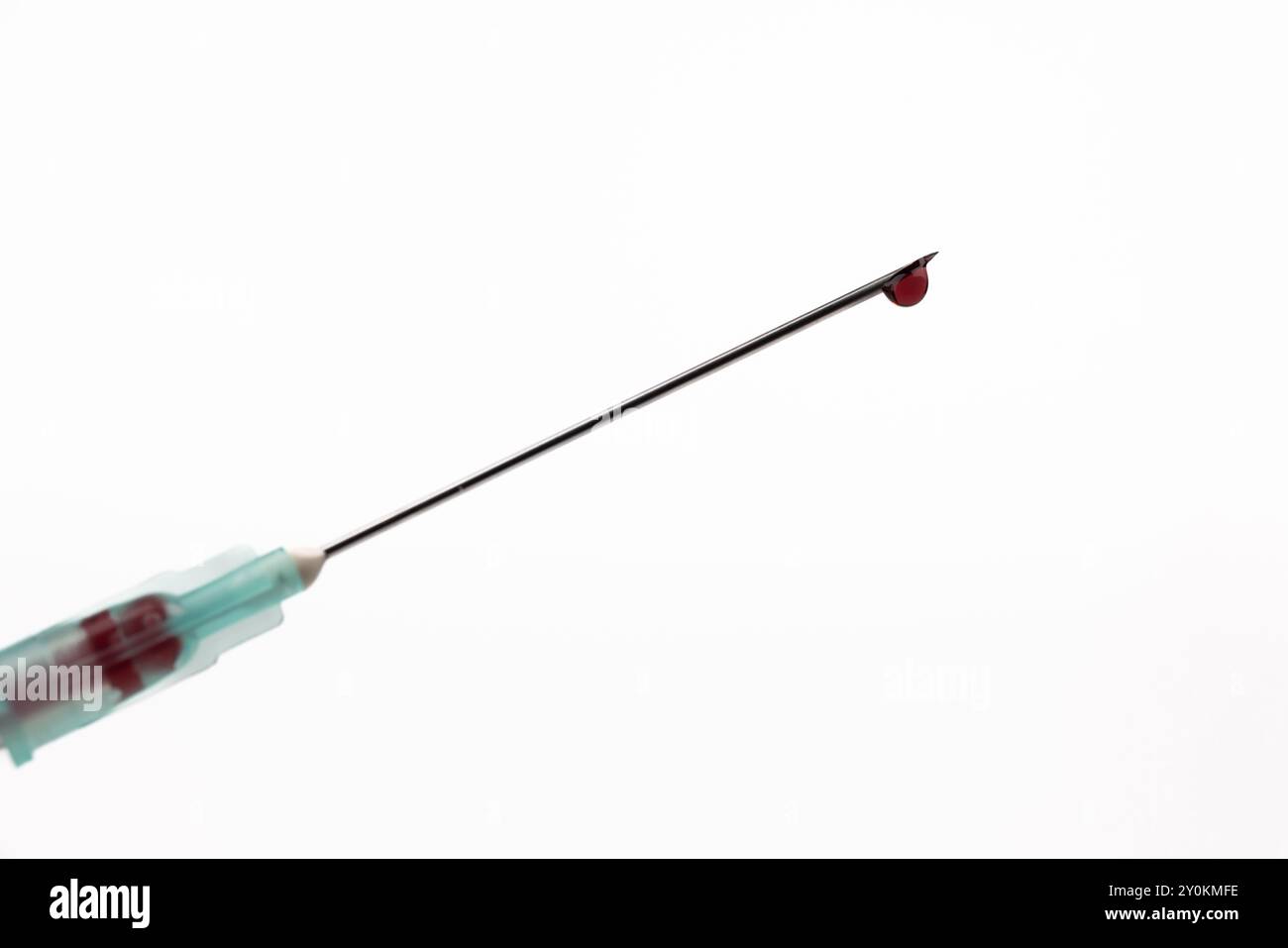A close-up of a syringe needle with a single drop of blood at its tip, centered in the horizontal frame. The image features a stark white background, Stock Photo