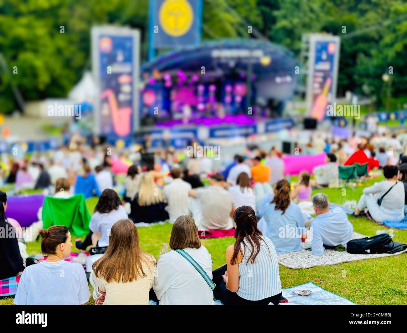 Large crowd gathered in park for outdoor music festival. Colorful blankets and chairs spread across grass while people enjoy live performance in sunlight. Stock Photo