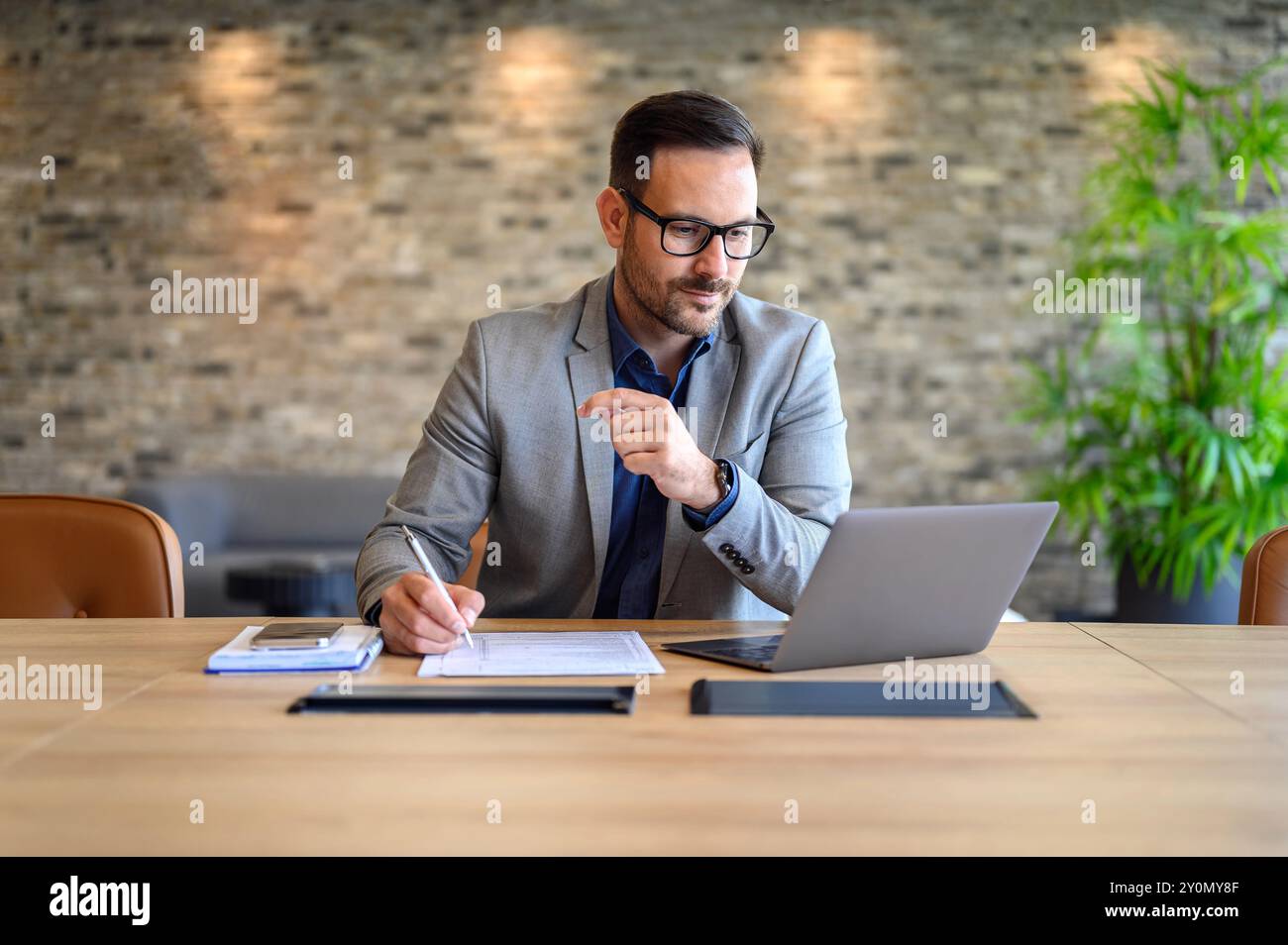 Focused male project manager writing on paper while doing online research over laptop at desk in office Stock Photo