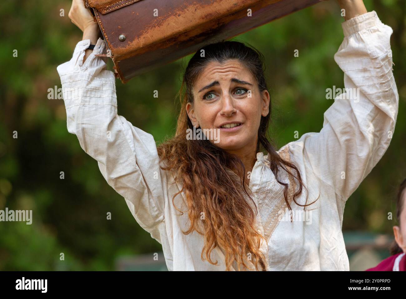 Actress in a white costume grimaces while lifting a heavy, vintage suitcase over her head during a theater performance Stock Photo