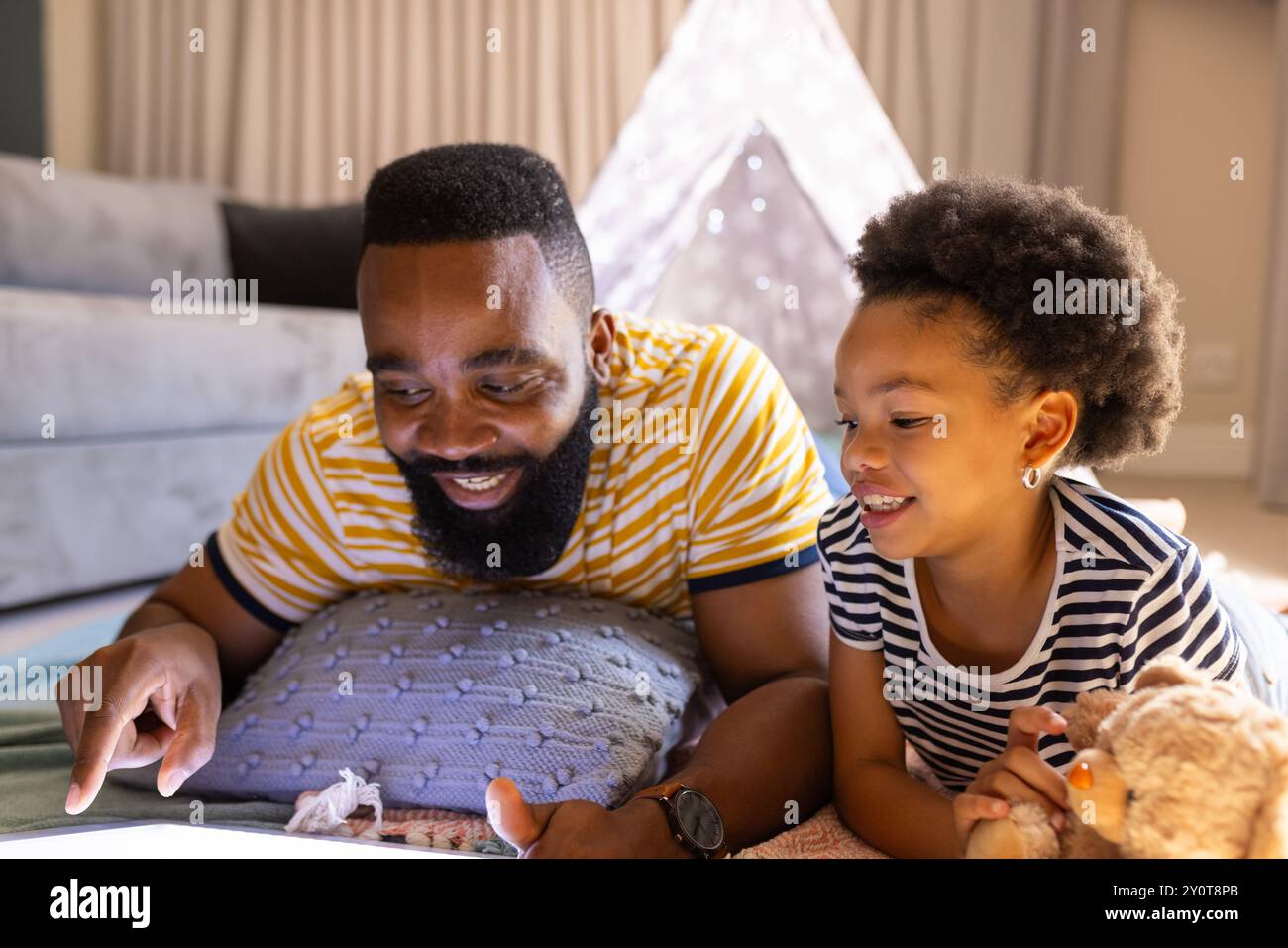 Father and daughter lying on floor, using tablet and smiling at home, in teepee Stock Photo