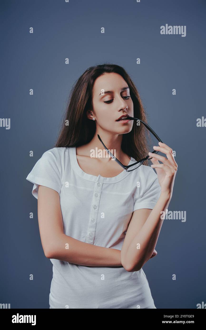 Young woman deep in thought, holding her eyeglasses in her mouth against a blue background. Suitable for education, business, and lifestyle concepts Stock Photo