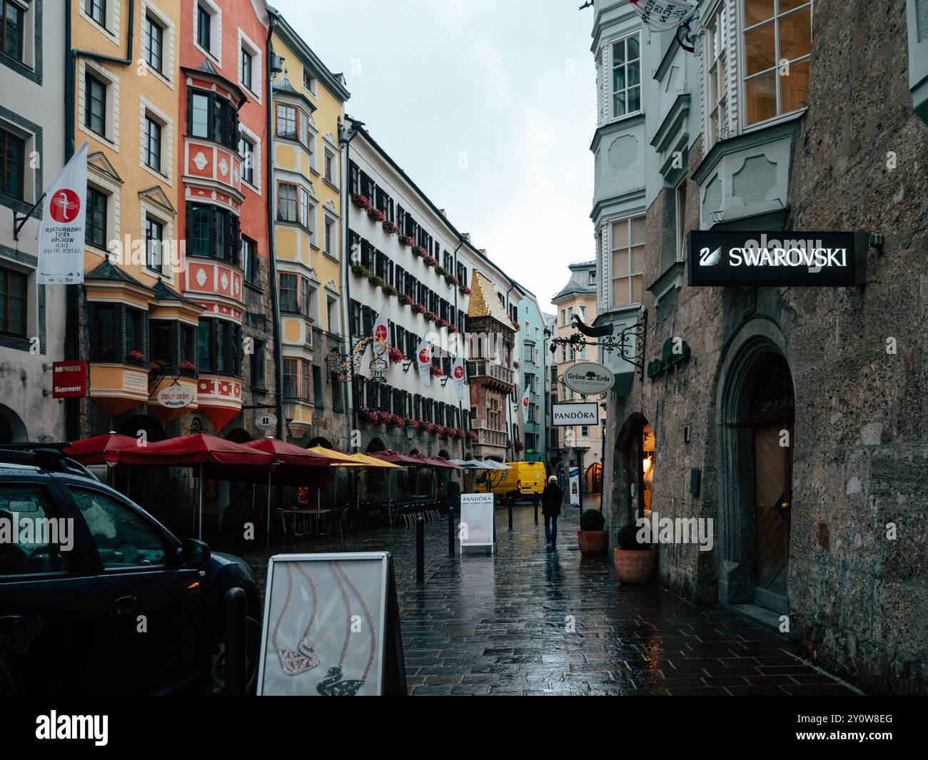 A quiet, rainy day in Innsbruck's city center features colorful buildings and shops. People stroll along the wet cobblestones, adding to the serene at Stock Photo