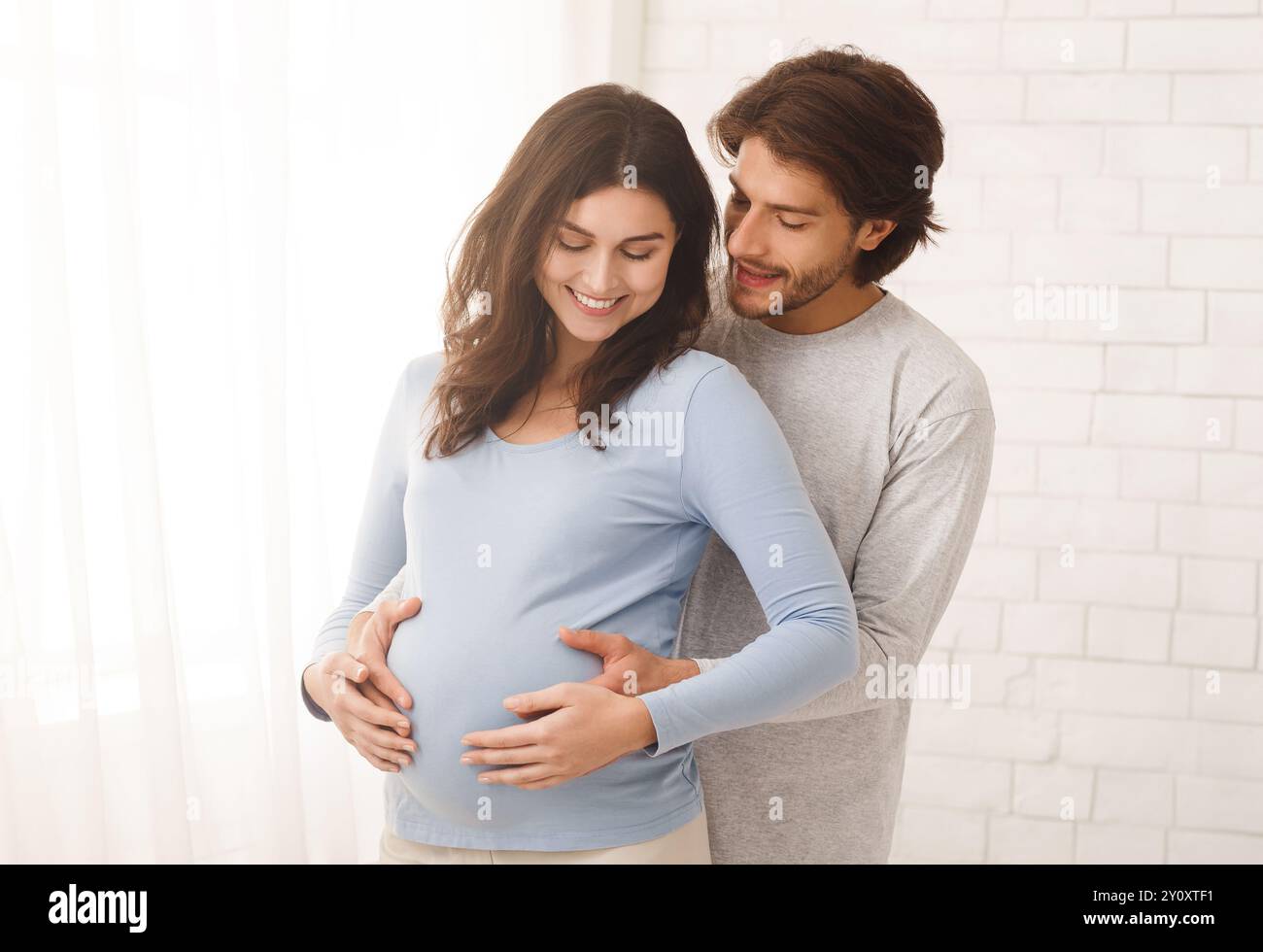 Loving man hugging his pregnant wife from behind standing near window ...