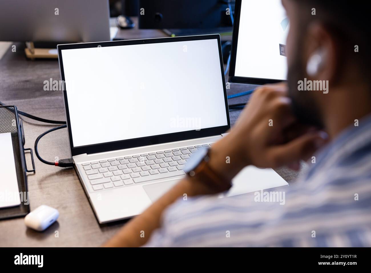 Using laptop with blank screen, Indian man thinking and wearing wireless earbuds, copy space Stock Photo