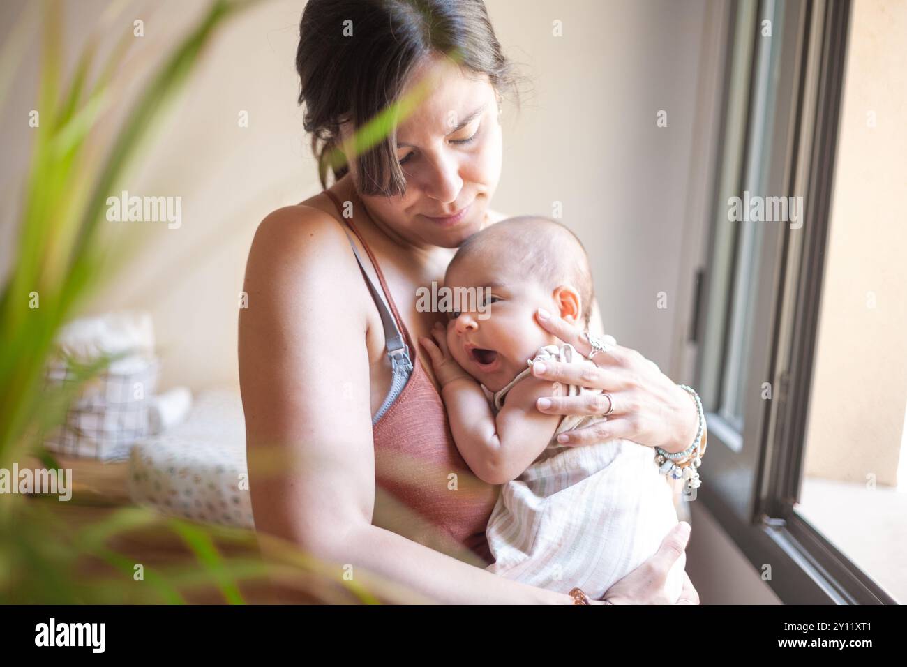 A woman holds a baby in her arms by a window. The woman looks at the baby and the scene conveys a sense of warmth and affection. Stock Photo