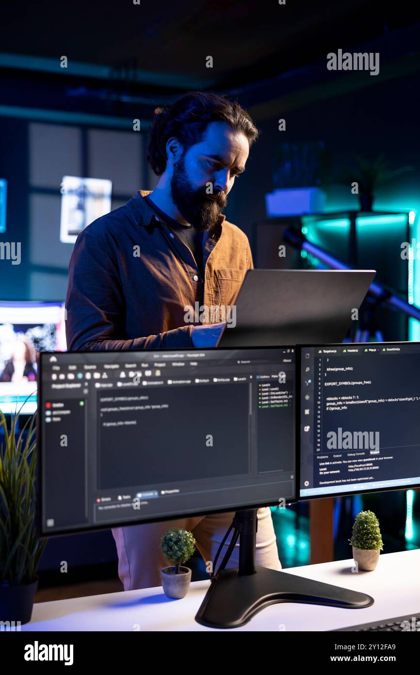 IT professional working from RGB lit home using laptop, testing and deploying programs and systems. Software engineer checking code displayed on computer monitors, standing in apartment Stock Photo