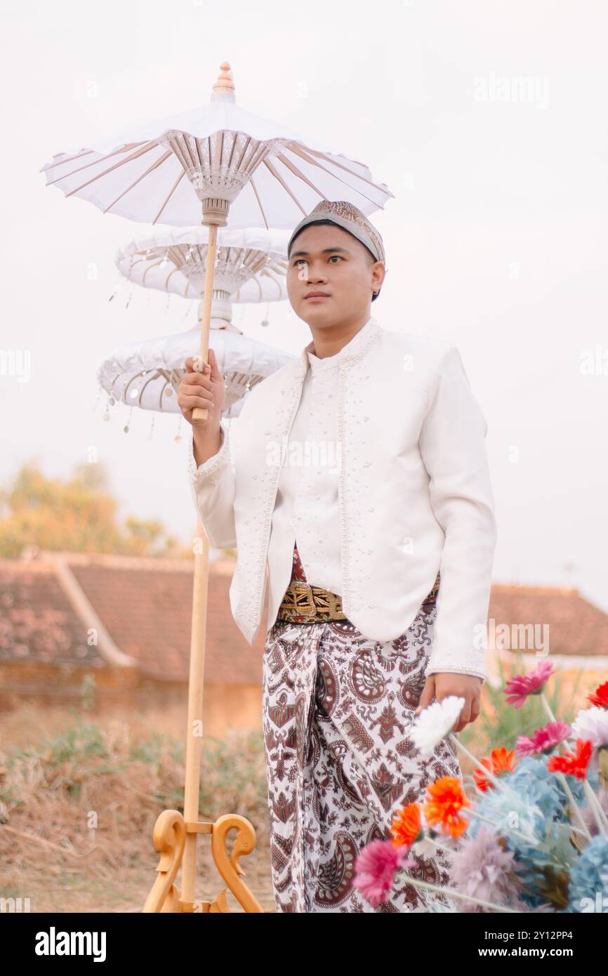 handsome young Asian male standing candidly holding an umbrella wearing modern Javanese traditional clothing, white velvet bridal kebaya in an outdoor Stock Photo