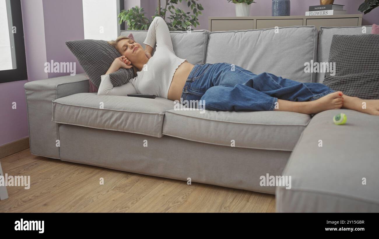 A young blonde woman relaxes on a sofa in a cozy, modern living room ...