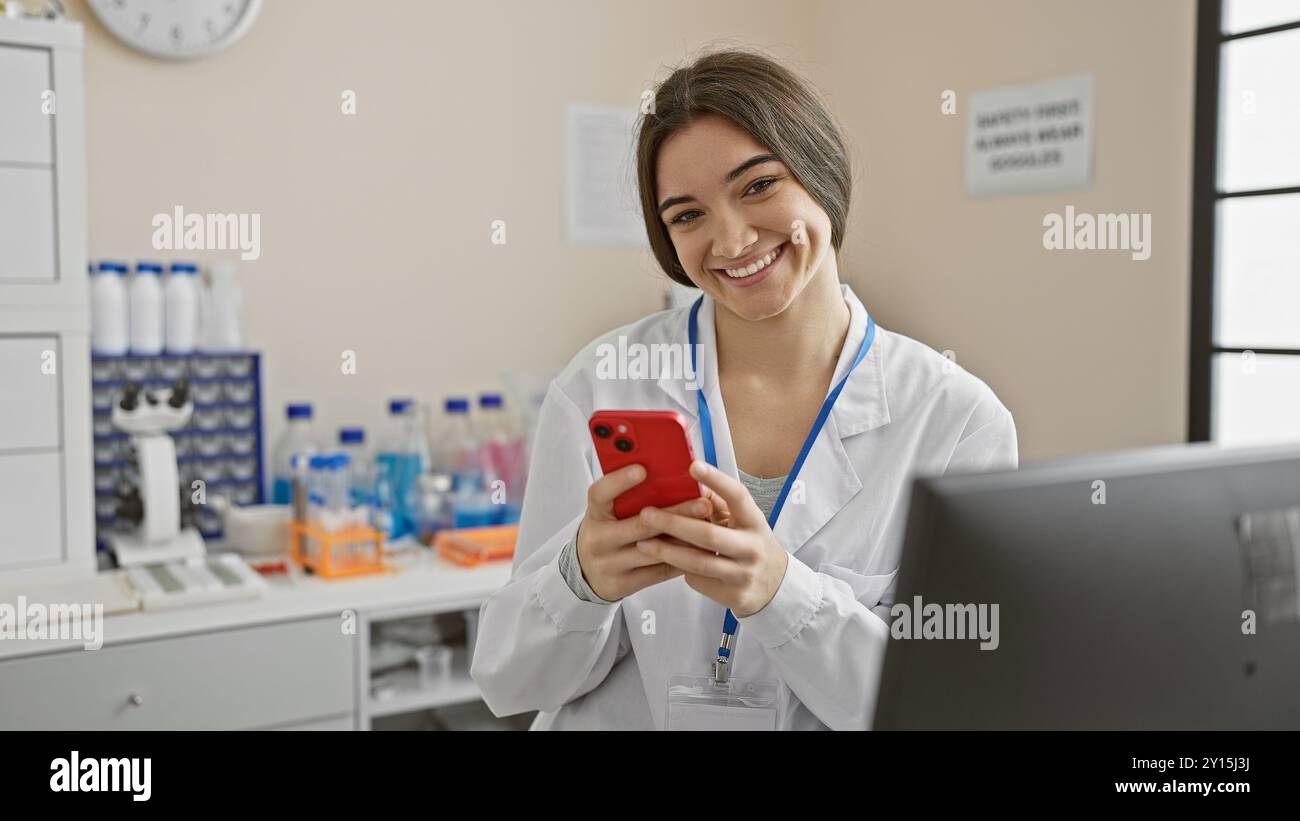 A smiling young hispanic woman in a lab coat using a smartphone indoors ...
