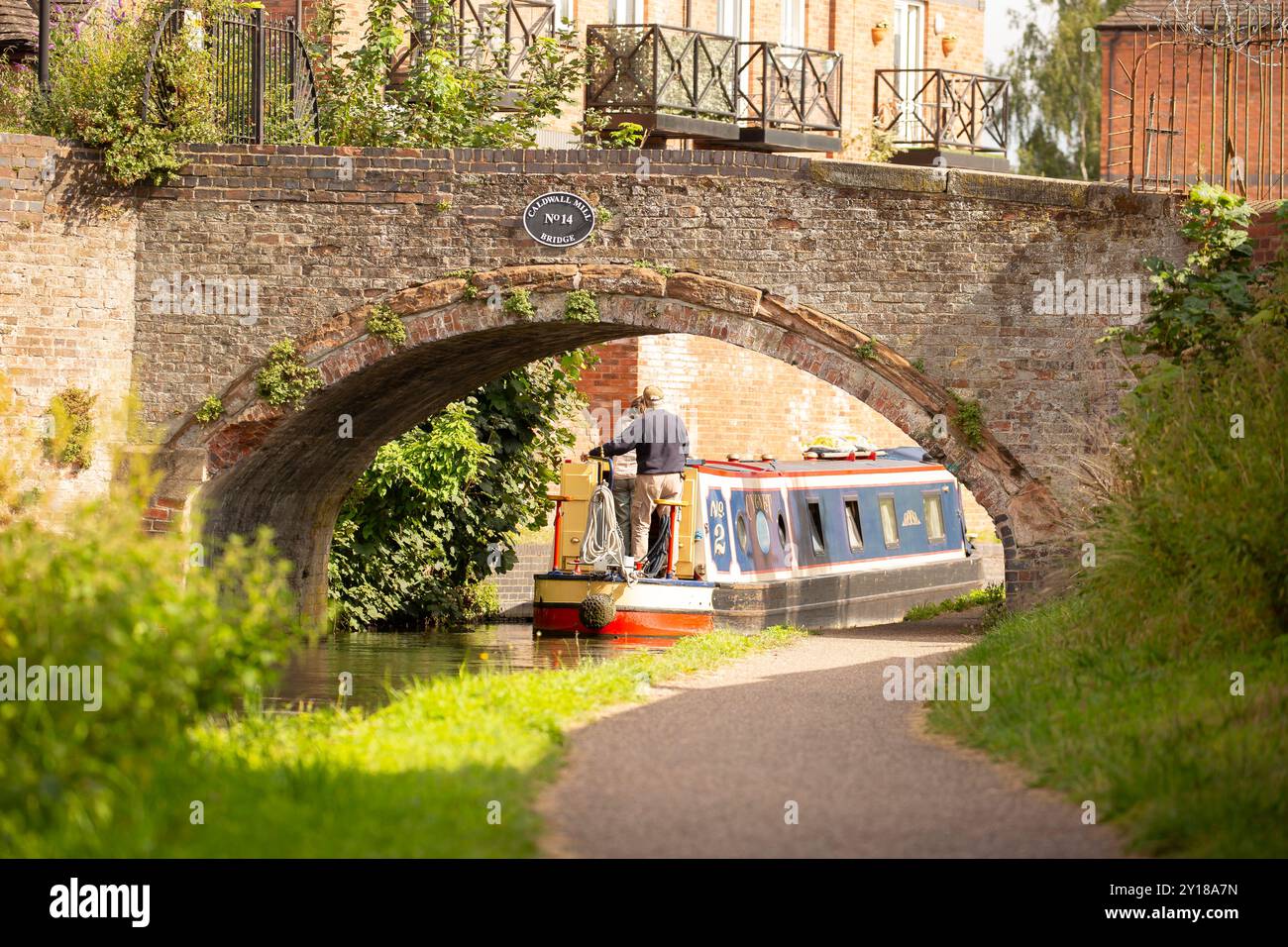 Rear view of couple on UK canal boat travelling under old brick arch bridge, properties with balconies in the background. Stock Photo