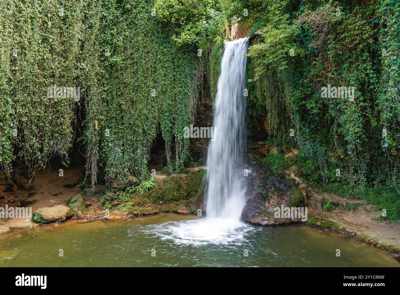 A serene waterfall cascades over rocks into a tranquil pool surrounded by lush greenery in a peaceful outdoor setting Stock Photo