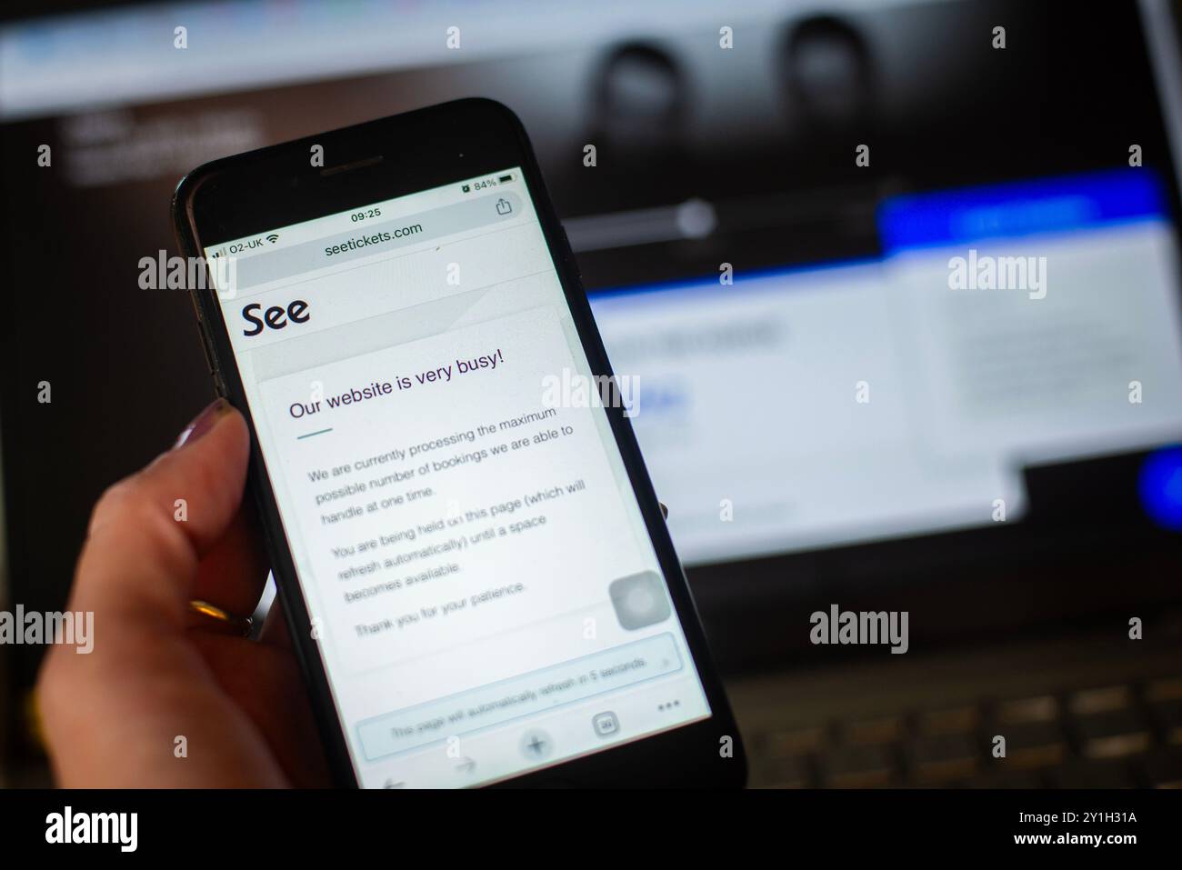 A hand holds a mobile phone screen showing See Tickets Website busy message for Oasis reunion tickets. Ticketmaster screen on laptop in background. Stock Photo