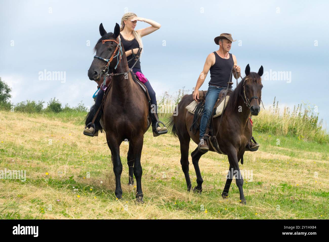 Two people on horseback riding through a grassy field, one looking into ...