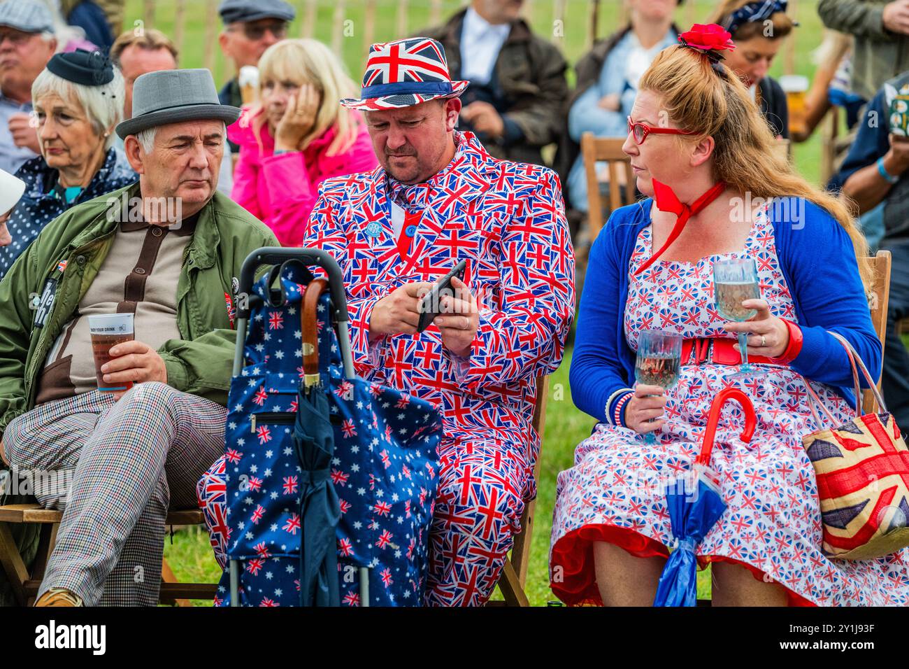 Goodwood, UK. 7th Sep, 2024. People take a breather in patriotic outfits, some with extra protection from the rain, which comes and goes - The Goodwood revival - The only historic motor race meeting to be staged entirely in a period theme, an immersive celebration of iconic cars and fashion. Credit: Guy Bell/Alamy Live News Stock Photo